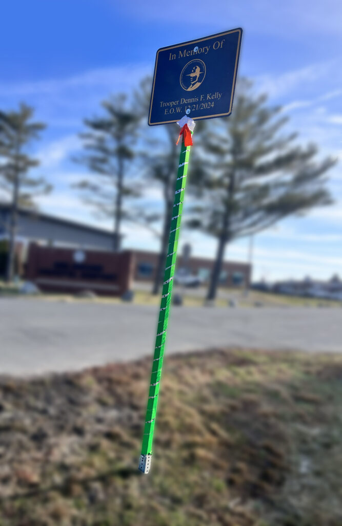 Blue sign the says "In Memory of Trooper Dennis F Kelly EOW 12/21/2024". Sign pole is wrapped in green ribbon with a white and orange bow. Background is blurred out building and blue sky.
